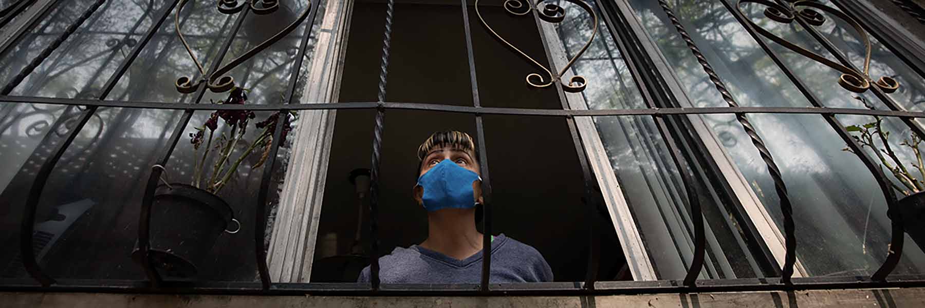 A person wearing a surgical mask while peering out of their window. Photo by Rodrigo Gonzalez on Unsplash.
